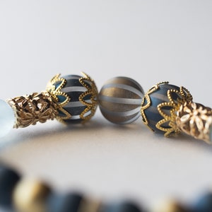 May include: Close-up of a beaded bracelet featuring gold-colored filigree accents and decorative beads. The bracelet includes dark blue and gold-striped spherical beads, and matte black beads. The design is elegant and detailed.