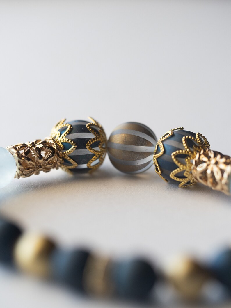 May include: Close-up of a beaded bracelet featuring gold-colored filigree accents and decorative beads. The bracelet includes dark blue and gold-striped spherical beads, and matte black beads. The design is elegant and detailed.