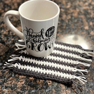 May include: White ceramic mug with black lettering that reads "Thank you for helping us GROW". The mug is sitting on a gray and white striped crocheted coaster.