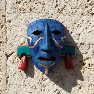May include: A blue mosaic mask with copper-colored accents and a turquoise and red side design. The mask has a prominent nose and eye openings, mounted on a textured, light-colored wall. The mask is a decorative art piece.