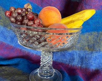 May include: A clear glass pedestal bowl with a decorative pattern, filled with red grapes, two yellow bananas, and two peach-colored fruits.