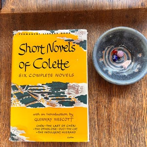 May include: A vintage book titled "Short Novels of Colette" with a yellow cover and green illustrations. The book is next to a small, speckled ceramic bowl containing colorful beads. The book's title and author are clearly visible.