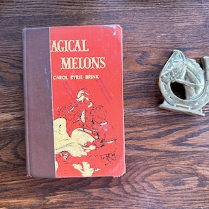 May include: A vintage book titled "Magical Melons" by Carol Ryrie Brink, featuring a red cover with illustrations and brown spine. A brass horseshoe-shaped object is next to the book, all on a wooden surface.
