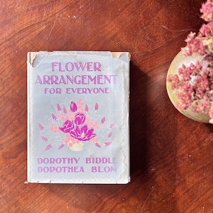 May include: A vintage book titled "Flower Arrangement for Everyone" with a light blue cover and pink floral illustrations. The book's cover features the authors' names, Dorothy Biddle and Dorothea Blom. The book is placed on a wooden surface.