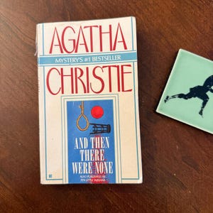 May include: A book cover for Agatha Christie's "And Then There Were None" with a blue and white design. The title is in red, and the cover features a noose and a red circle. A small, square, light green tile with a black silhouette is to the right.
