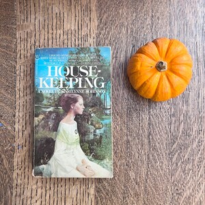 May include: A vintage paperback book titled "Housekeeping" by Marilynne Robinson, featuring a woman in a white dress on the cover. Beside the book is a small, bright orange pumpkin.