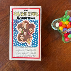 May include: A vintage paperback book titled "The Rolling Stone Interviews" with a blue star-patterned background and portraits of musicians. The book is next to a green glass dish filled with colorful jelly beans.
