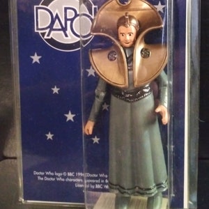 May include: A plastic blister pack containing a Doctor Who action figure. The figure is wearing a grey robe and a gold helmet. The figure is from the 1996 Doctor Who series. The packaging is blue with white stars and the text "Doctor Who" in large, silver letters.
