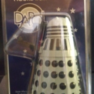 May include: A white plastic Doctor Who Dalek figure from the BBC television series. The figure is packaged in a clear plastic blister pack with a blue and white background. The packaging features the Doctor Who logo and the text "FIGURE from"
