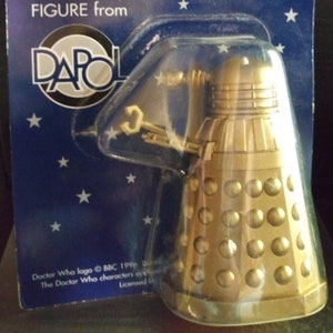 May include: A gold-coloured Doctor Who Dalek figure from the Dapol brand. The figure is packaged in a blue and white cardboard box with stars. The box has the text "Another Famous Doctor Who Figure from Dapol" on it.