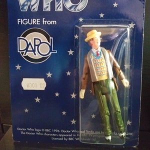 May include: A blue and white card with the words "Another Famous Doctor Who Figure from DAPO" and a picture of a plastic action figure of the Doctor from the British science fiction television series Doctor Who. The figure is wearing a brown jacket, a yellow sweater with a question mark pattern, green trousers, and a yellow hat. The card also has the number "WO01 02" printed on it.