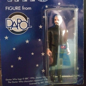 May include: A Doctor Who action figure from DAPOL, packaged on a blue card with white stars. The figure is of a man in a black coat and trousers, with a grey beard and hair. The packaging reads "ANOTHER DOCTOR WHO FIGURE from DAPOL."