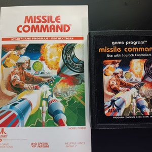 May include: A vintage Atari game cartridge for Missile Command. The cartridge is black with a colorful image of a spaceship and a city under attack. The text "game program™ missile command™ Use with Joystick Controllers" is printed on the front of the cartridge.