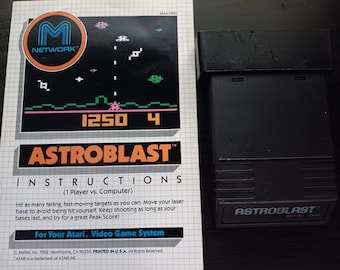 May include: A black Atari video game cartridge with the title "ASTROBLAST" printed on the label. The cartridge is next to a printed instruction sheet with a screenshot of the game and the text "ASTROBLAST" in orange.