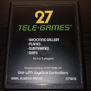 May include: A black Atari 2600 game cartridge with a yellow number 27 and the text "TELE-GAMES®". The cartridge has a list of games: "SHOOTING GALLERY", "PLANES", "SUBMARINES", and "SHIPS". The text "for 1 or 2 players" is also on the cartridge. The text "Program contents © 1977 ATARI, INC." and "Use with Joystick Controllers" is printed on the cartridge. The text "SEARS, ROEBUCK AND CO." and "4975102" is also printed on the cartridge.
