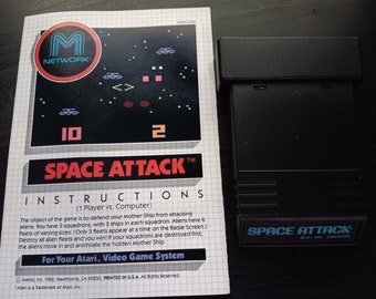 Puede incluir: Un cartucho de videojuego Atari negro con una etiqueta azul que dice "SPACE ATTACK" y "M.I. 1982 SINGAPORE". El cartucho está al lado de un folleto de instrucciones en blanco y negro con el título "SPACE ATTACK" y una pequeña imagen del juego.