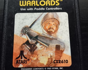 May include: A black Atari game cartridge with a yellow label. The label reads "game program™ WARLORDS™ Use with Paddle Controllers" and features a knight holding a sword. The cartridge is labeled "ATARI" and "CX2610". The text "PROGRAM CONTENTS © 1981 ATARI, INC." is printed at the bottom of the label.