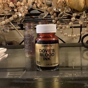 May include: A small glass bottle of "Dove's Blood Ink" with a white cap, next to a smaller bottle labeled "Witches & Brooms." The ink bottle is 1 fl oz (29.5ml). The background includes floral decorations.