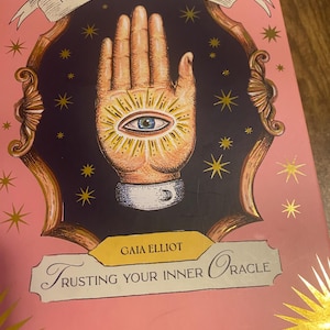 May include: A pink book titled "The Book of Answers" with gold accents. The cover features a detailed illustration of a hand with an eye in the palm, surrounded by radiating lines and stars. The author's name is "Gaia Elliot".