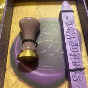 Puede incluir: Un sello de cera morado y una barra de cera a juego se muestran dentro de una caja decorativa. El sello tiene una base dorada y un mango morado. La barra de cera es morada y tiene la inscripción "Sealing Wax".