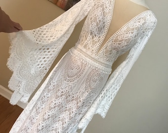 May include: A white lace wedding dress with long sleeves and a plunging neckline. The dress has a fitted bodice and a flowing skirt. The sleeves are bell-shaped and have a delicate lace trim.