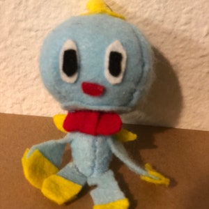 May include: A blue and yellow plush toy with a red bow. The toy has a round head with large eyes and a small mouth. The toy is sitting on a brown surface.