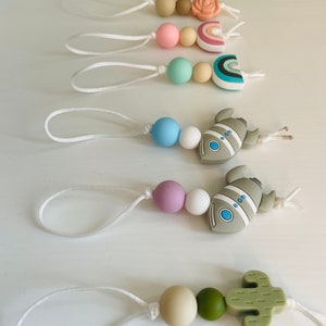 Può includere: Cinque morsi di silicone con diversi design e colori. Ogni morso ha una perla bianca e una corda bianca attaccata. I design includono un fiore, un arcobaleno, un razzo, un razzo e un cactus.