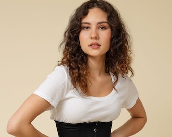 Puede incluir: Un corsé negro con una camiseta blanca y una falda verde. El corsé tiene una serie de ganchos y ojales metálicos para el cierre.