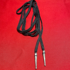 Puede incluir: Cordones negros para zapatos con puntas de metal plateadas. Los cordones están enrollados y colocados sobre una superficie roja.