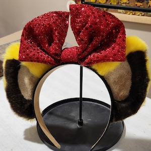 May include: A headband featuring plush, animal-like ears in yellow, brown, and black, topped with a large, sparkly red bow. The headband is displayed on a black stand, showcasing the playful design. The ears have a fluffy texture, adding to the whimsical aesthetic.