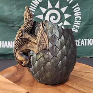 May include: A decorative, dragon-themed planter. The planter is shaped like a dragon egg, with a detailed, textured surface. A golden-brown dragon is sculpted on the side. The planter is set on a wooden surface.