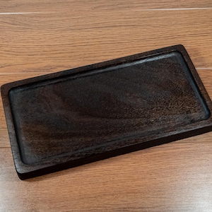 May include: A rectangular, dark brown wooden tray with a recessed interior. The tray has a raised border and is made of a wood with a visible grain pattern. The tray is displayed on a light brown wooden surface.