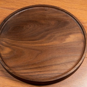May include: A round wooden serving tray made of dark walnut wood. The tray has a smooth, polished surface and a slightly raised edge.