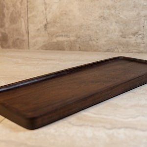 Puede incluir: Una bandeja de madera rectangular larga y estrecha, hecha de madera de nogal oscuro. La bandeja tiene un borde ligeramente elevado y una superficie lisa y pulida.