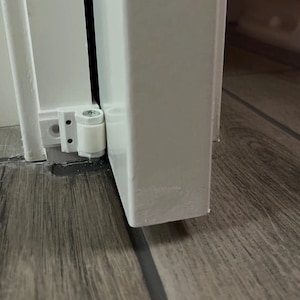 May include: Close-up of a white door and frame with a white hinge and a white cylindrical door stop. The door and frame are set against a wood-look floor.