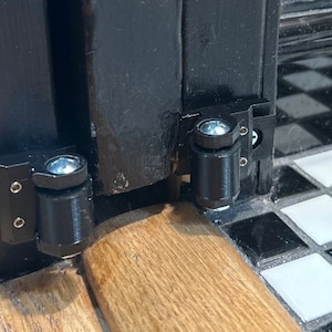 May include: Close-up of two black door hinges with silver screws. The hinges are attached to a black door frame and a wooden floor.