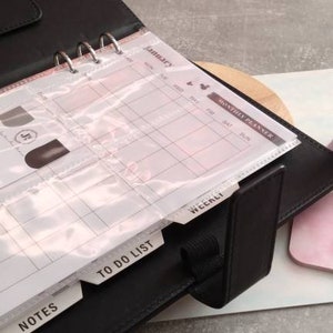 May include: A black leather bound planner with a clear plastic insert for a weekly calendar. The planner has tabs for "Notes", "To Do List", and "Weekly".