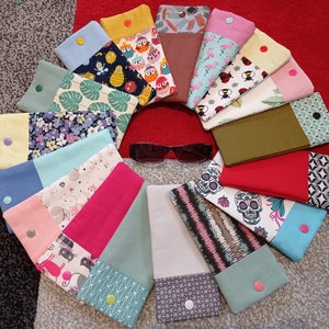 Peut inclure: Une collection de 15 étuis à lunettes en tissu coloré avec différents motifs et designs. Les étuis sont disposés en cercle avec une paire de lunettes de soleil au centre.