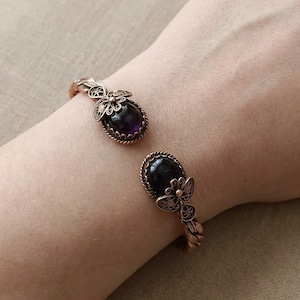 Può includere: Un bracciale a fascia in rame con due estremità aperte, ciascuna decorata con una pietra preziosa ovale viola scuro incastonata in una cornice in filigrana. La fascia metallica ha una trama a corda intrecciata e la filigrana ricorda un motivo floreale.