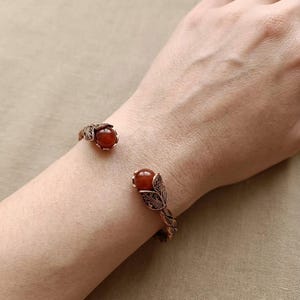 Peut inclure: Un bracelet manchette ouvert de couleur cuivre, orné à chaque extrémité d'une perle en pierre semi-précieuse ronde et polie de couleur brun rougeâtre. La bande métallique présente un motif complexe en relief rappelant des feuilles.