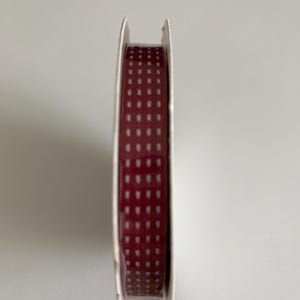 May include: A spool of narrow ribbon featuring a deep burgundy red base color with a repeating pattern of small, white, rectangular dashes arranged in vertical columns along the entire length of the fabric material.