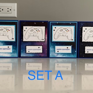 May include: Set of six blue and white birthday cards with a galaxy background and a white video game controller graphic. Each card has the text "Happy Birthday" printed on it.