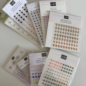 May include: A collection of Stampin' Up! adhesive-backed embellishments in various shapes and colors, including pearls, enamel pebbles, hexagons, and flower shapes, packaged in clear plastic sleeves with white header cards featuring brand text.