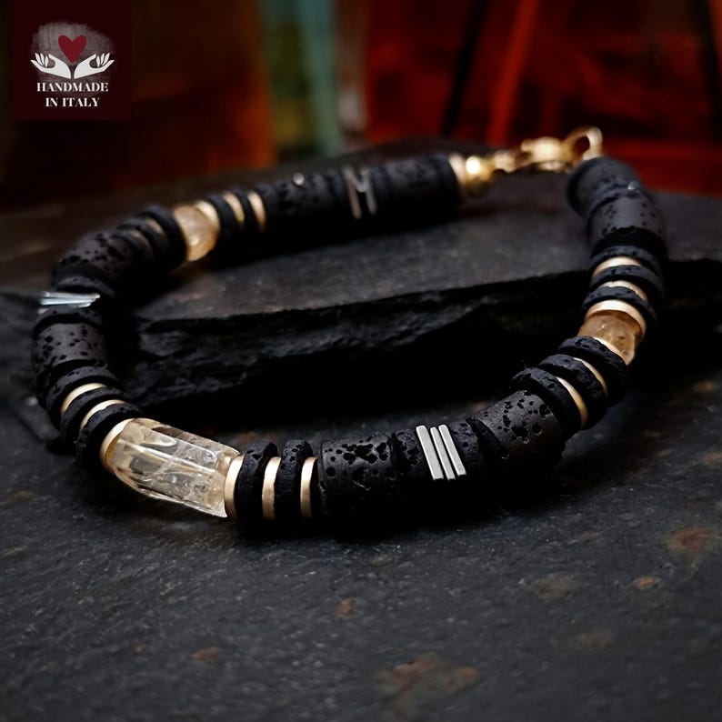 May include: Black lava rock bracelet with gold-colored accents and clear, rectangular crystal beads. The bracelet features silver-colored metal bars and the text "HANDMADE IN ITALY" with a heart logo.