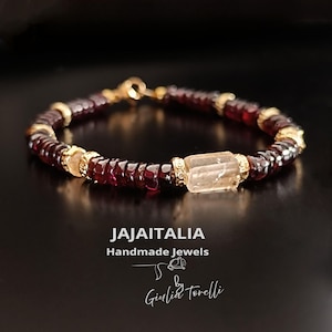 Op de afbeelding: Een handgemaakte armband met donkerrode kralen en gouden accenten. De armband heeft een rechthoekige, lichtgekleurde steen in het midden. De tekst "JAJAITALIA Handmade Jewels" en een handtekening zijn zichtbaar.