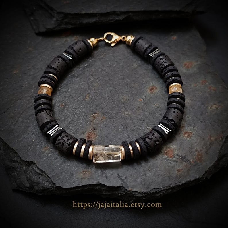 May include: A black beaded bracelet with gold accents and a rectangular, clear stone centerpiece. The bracelet features dark gray, circular beads, gold-colored spacers, and a gold clasp. The Etsy shop URL is visible at the bottom.