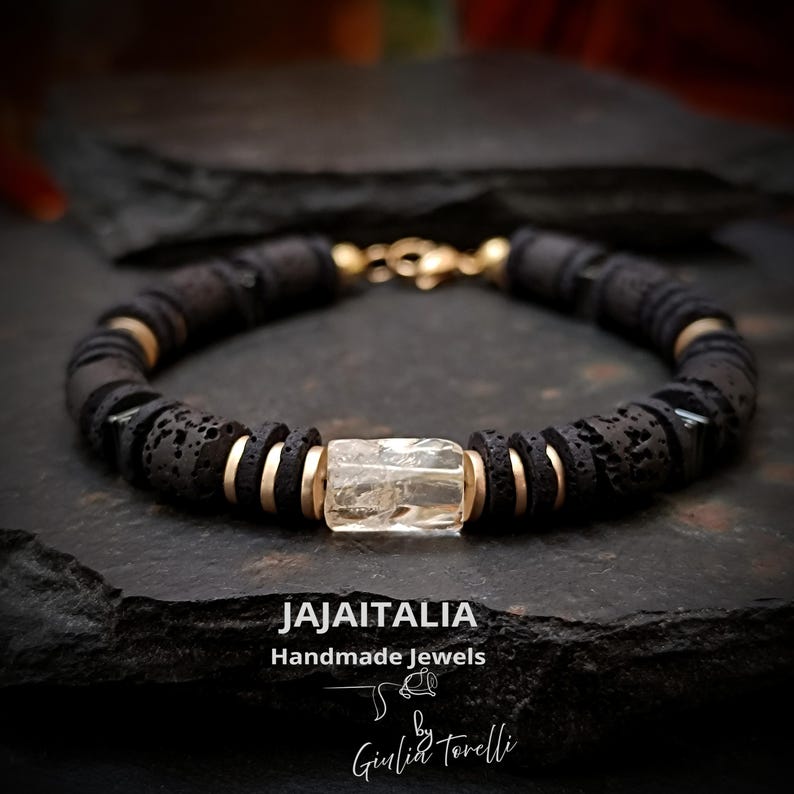 May include: A black lava stone bracelet with gold-colored accents and a clear, rectangular gemstone. The bracelet is handmade and features a gold clasp. The brand name "JAJAITALIA" and "Handmade Jewels" are visible.