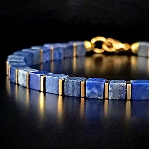 以下が含まれることがあります： 正方形の濃い青色のビーズと金色のアクセントが特徴のブレスレットのクローズアップ。ブレスレットは金色の留め金で留められています。ビーズのサイズは約0.6cmです。背景は黒です。
