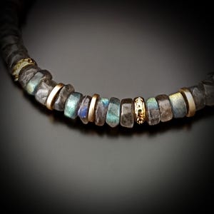K&ouml;nnte beinhalten: Halskette mit grauen, irisierenden Labradoritperlen, abwechselnd mit goldfarbenen Metallringen. Der dunkle Hintergrund hebt die Farben der Steine und des Metalls hervor.