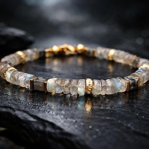 May include: A beaded bracelet featuring iridescent, rectangular and round-cut stones in shades of gray, blue, and clear. The bracelet is accented with gold-colored metal spacers and a clasp. The jewelry is displayed on a dark, textured surface.